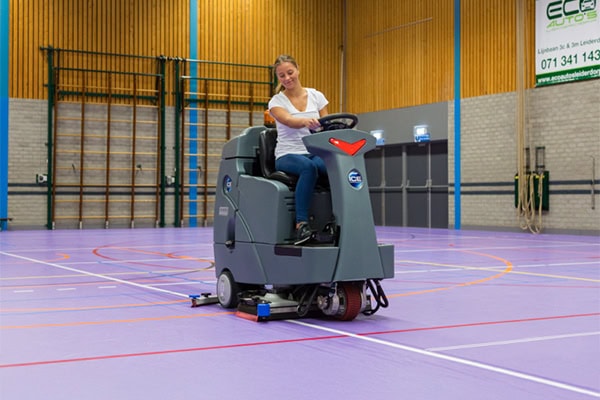 Mopmachine voor vloerenreiniging in sporthal met vrouwelijke bestuurder, benadrukt innovatieve schoonmaaktechnologie door ICE Cobotics.